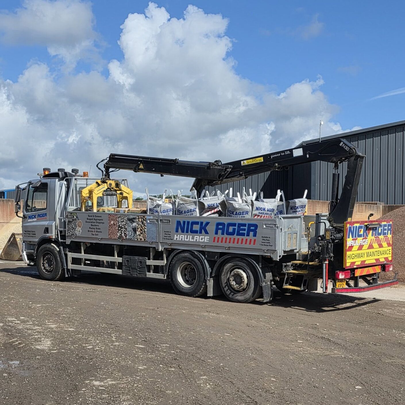 A Nick Ager Haulage truck equipped with a crane, loaded with bags of materials, parked at a construction site under a blue sky with fluffy clouds.