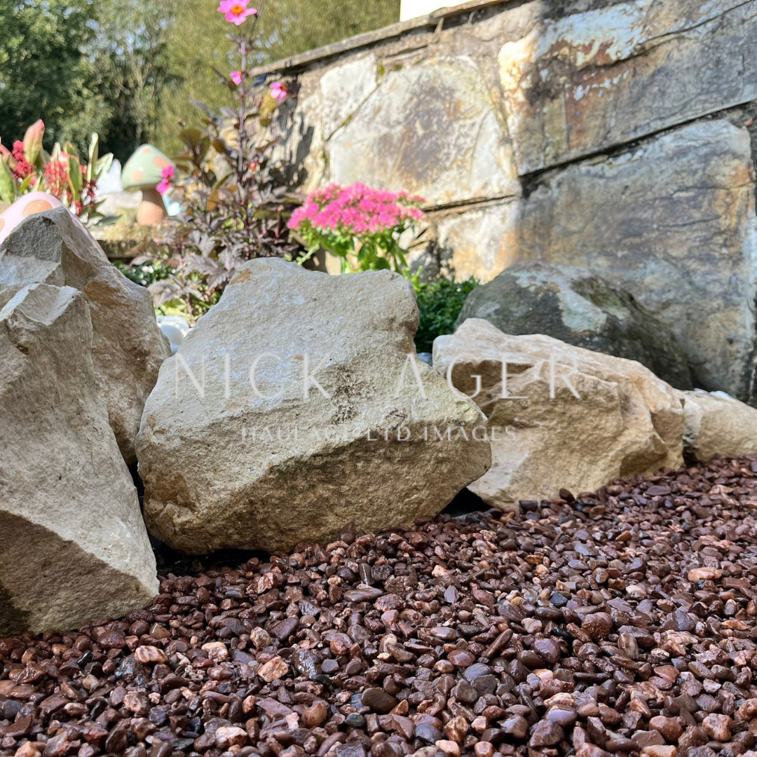 A close-up view of large decorative rocks surrounded by brown gravel, with colourful flowers in the background and a stone wall visible.