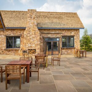 A stone house featuring a wooden roof, outdoor wooden furniture, and a landscaped area with trees.