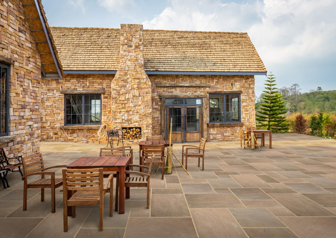 A stone house featuring a wooden roof, outdoor wooden furniture, and a landscaped area with trees.
