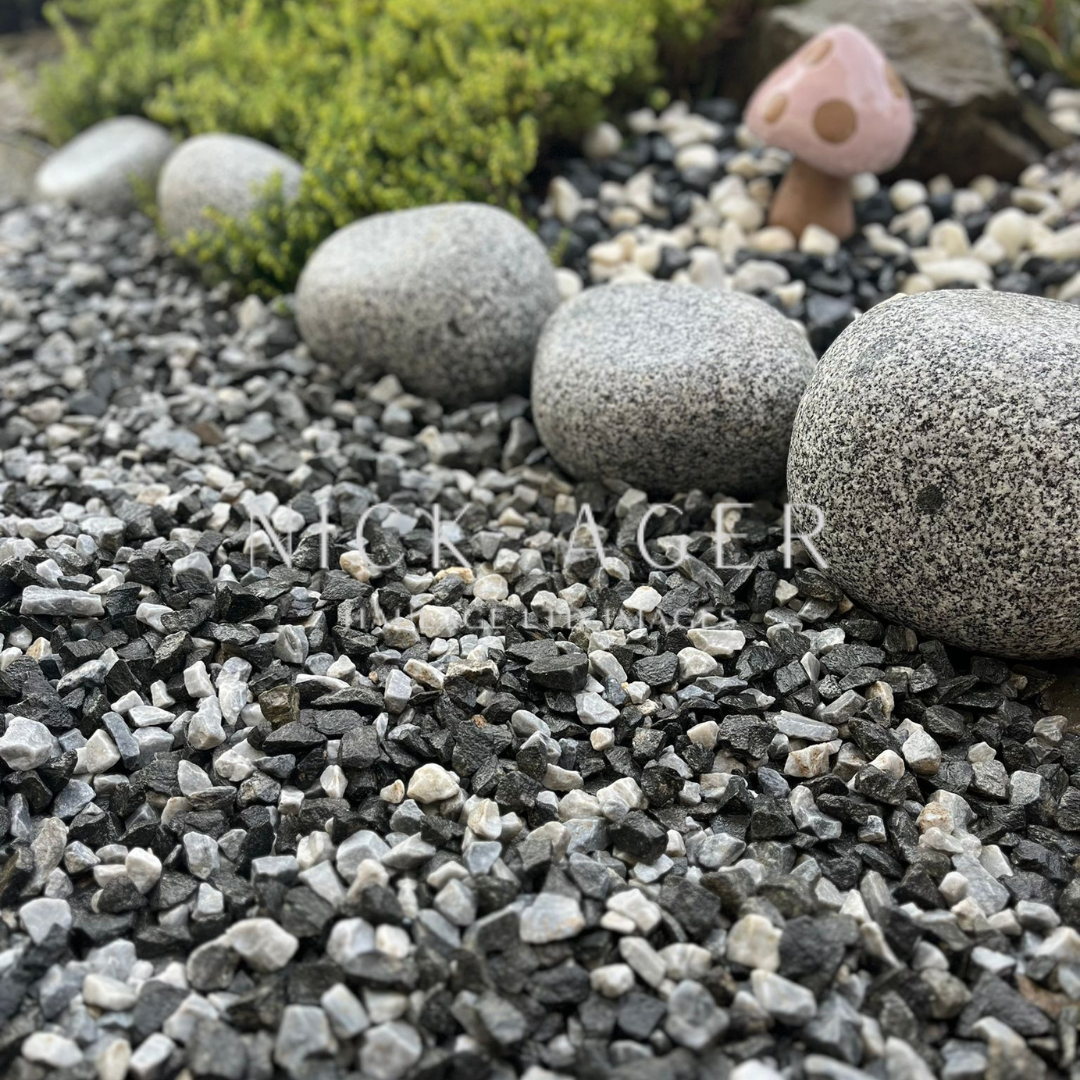 A close-up view of decorative gravel featuring black, grey, and white stones, alongside larger smooth grey rocks and a small pink mushroom ornament in a garden setting.