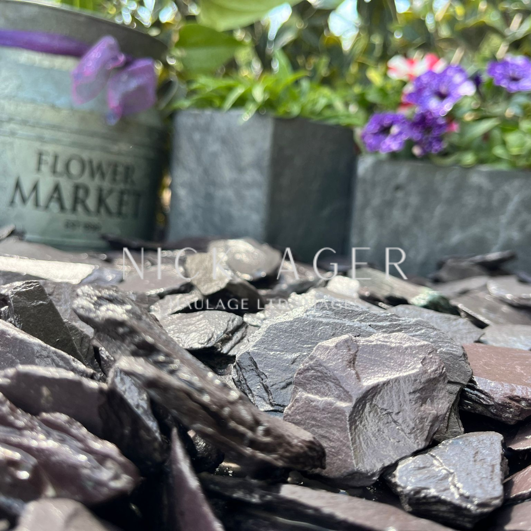 A close-up view of decorative stones in various shades of grey and purple, with plant containers in the background, including a metal Flower Market pot and two grey stone pots holding greenery and flowers.