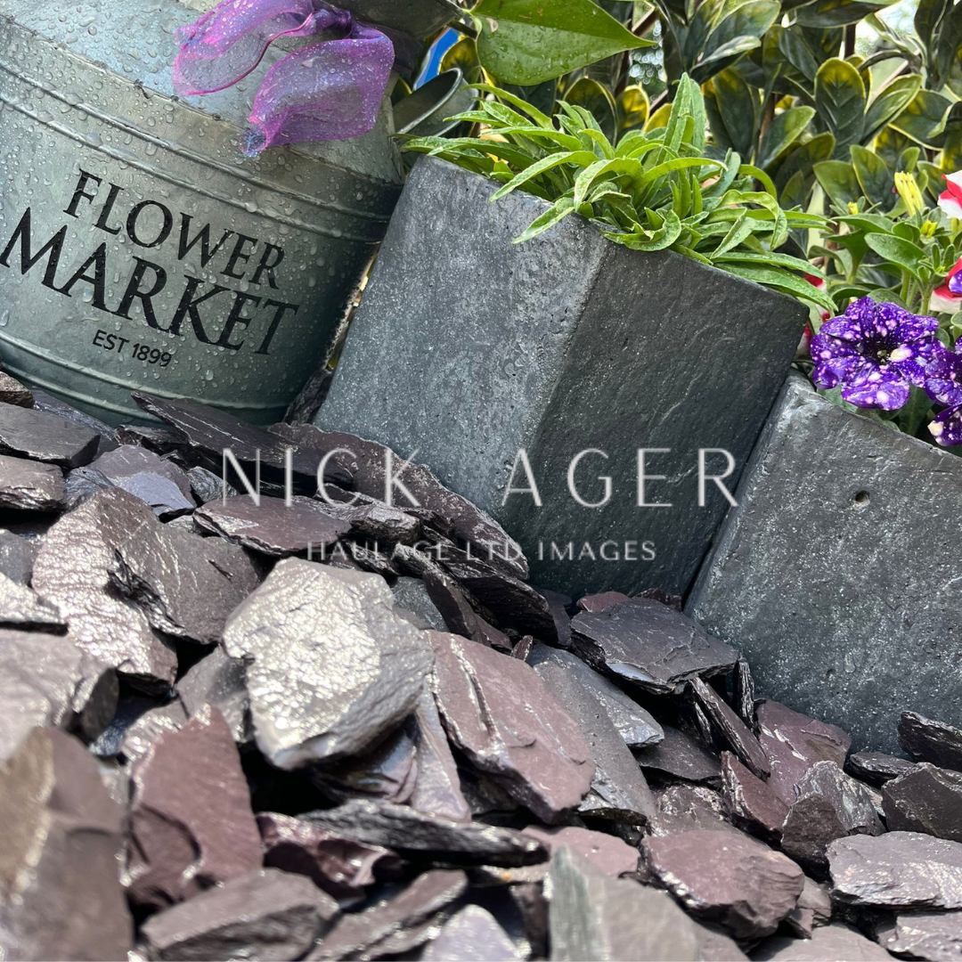 A decorative arrangement featuring a galvanized tin pot labelled 'Flower Market' with plants, two rectangular stone planters, and a bed of dark slate stones.
