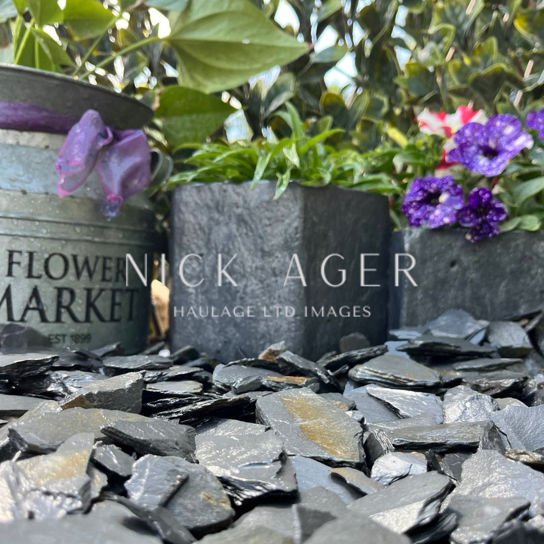 A close-up view of a flower market scene featuring a silver metal pot, a dark stone planter with green plants, and purple flowers, all situated on a bed of black decorative gravel.