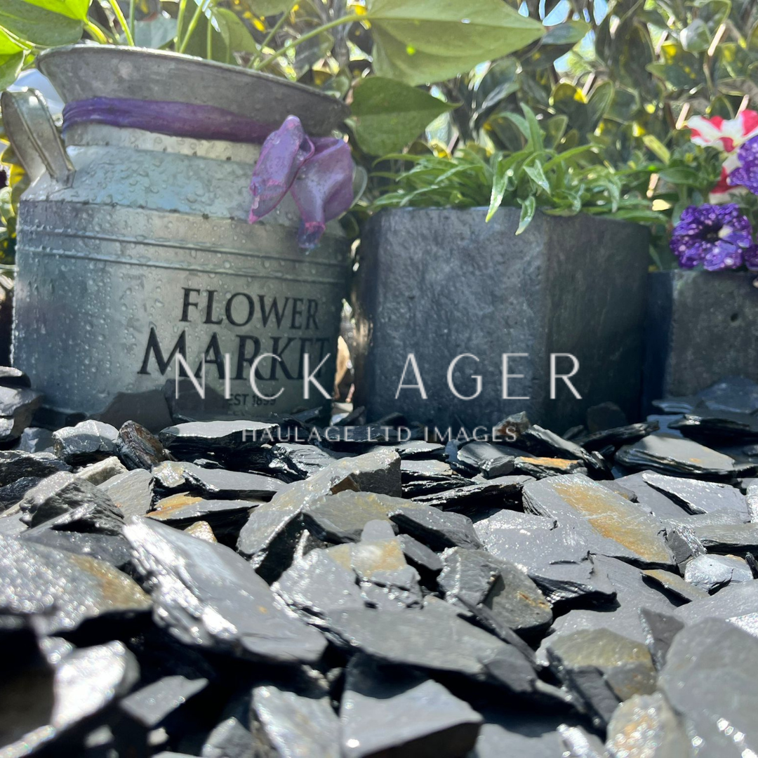 A rustic metal container labelled 'Flower Market' sits alongside a square stone planter, surrounded by vibrant flowers and black slate pebbles.