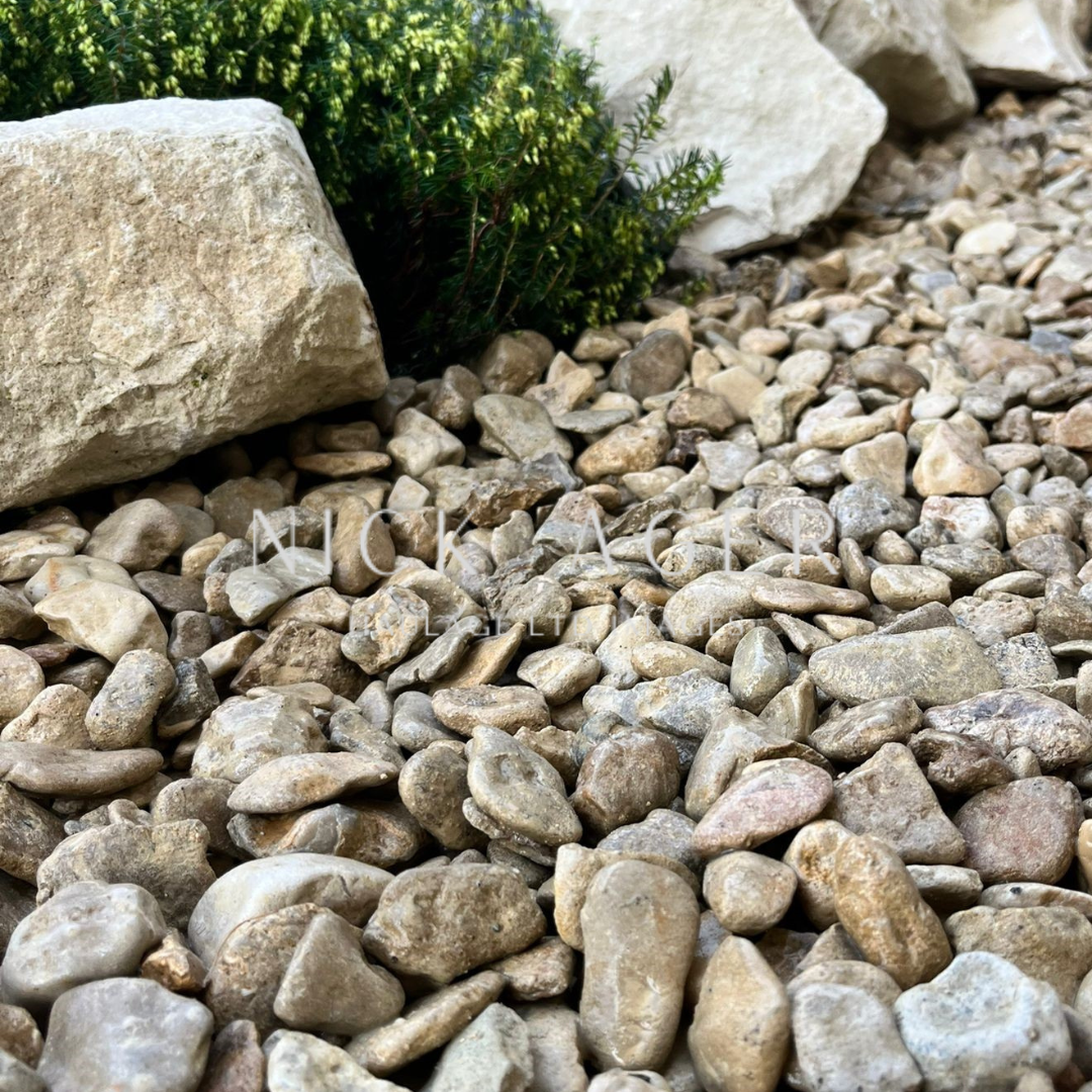 Close-up of naturally rounded gravel stones in varying shades of beige and brown, with a background of greenery and large rocks.