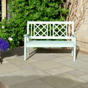 A mint green wooden bench with a lattice back rests on a stone paved patio, surrounded by colourful flowering plants and greenery.