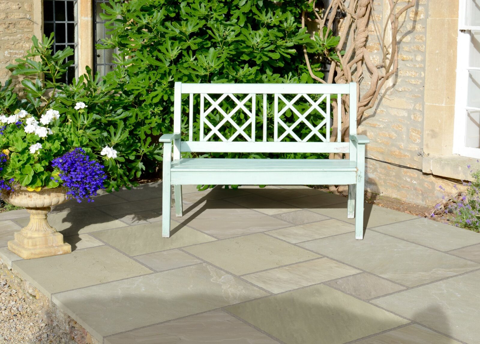 A mint green wooden bench with a lattice back rests on a stone paved patio, surrounded by colourful flowering plants and greenery.