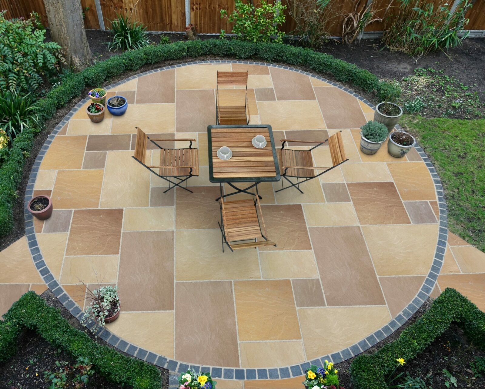 An overhead view of a circular patio area with light and dark stone paving, featuring a wooden table and four folding chairs. Surrounded by neatly trimmed hedges and various potted plants.