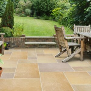 A picturesque garden patio featuring wooden seating, a dining table, and potted sunflowers. The stone paving leads to a lush green lawn surrounded by abundant greenery.