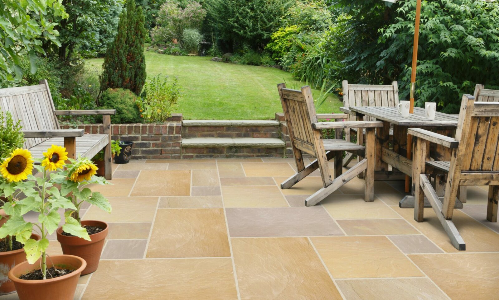 A picturesque garden patio featuring wooden seating, a dining table, and potted sunflowers. The stone paving leads to a lush green lawn surrounded by abundant greenery.