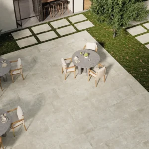 Aerial view of a contemporary outdoor dining space featuring two round tables with wooden chairs, surrounded by grey stone flooring and green grass, and a backdrop of a modern building.