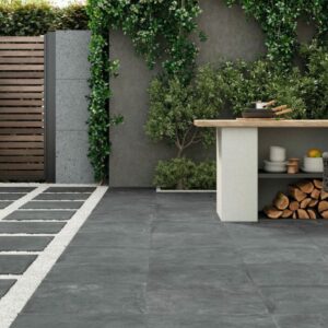 A contemporary garden patio featuring grey tiled flooring, a wooden dining table with wicker chairs, an outdoor kitchen area, and various plants in decorative pots.