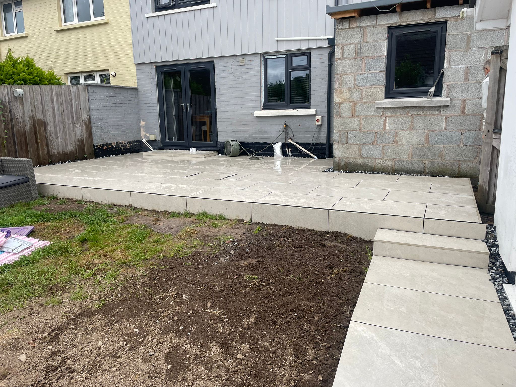 A newly installed outdoor patio featuring large grey paving stones, a green lawn area, and an adjacent building with grey brick walls.