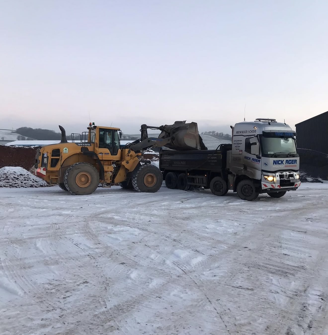 Heavy Machinery Loading Snowy Ground A yellow Volvo wheel loader scooping material next to a grey truck in a snowy landscape with hills in the background.