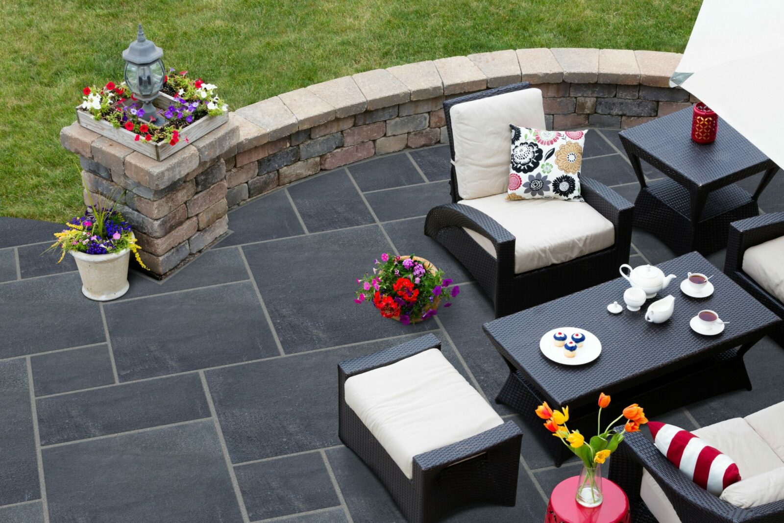 A beautifully arranged outdoor patio featuring wicker chairs, a coffee table with tea and pastries, and vibrant flower pots.