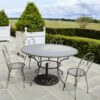 A round grey outdoor dining table accompanied by four metal chairs, set on a stone patio with well-trimmed hedges in the background.