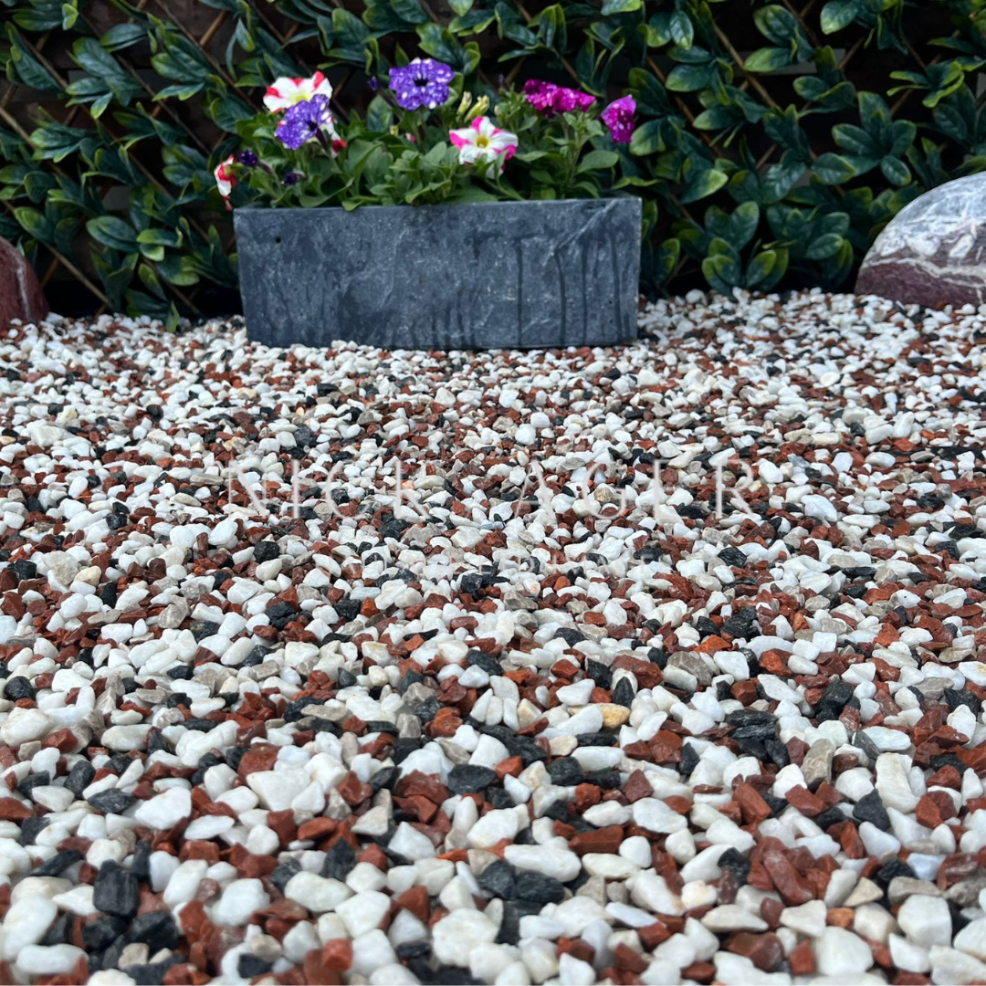 A close-up view of multicoloured decorative gravel, featuring red, black, and white stones, with a dark stone planter filled with blooming purple and pink flowers in the background.