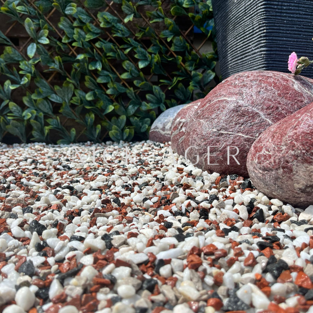 A close-up view of a garden area featuring decorative gravel with white, red, and black stones, complemented by larger pink and grey rocks and lush green foliage in the background.