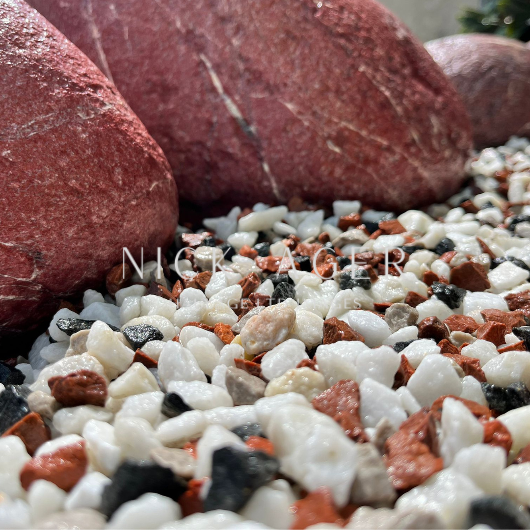 A close-up view of smooth red rocks resting on a bed of small, multi-coloured pebbles including white, black, and reddish tones.