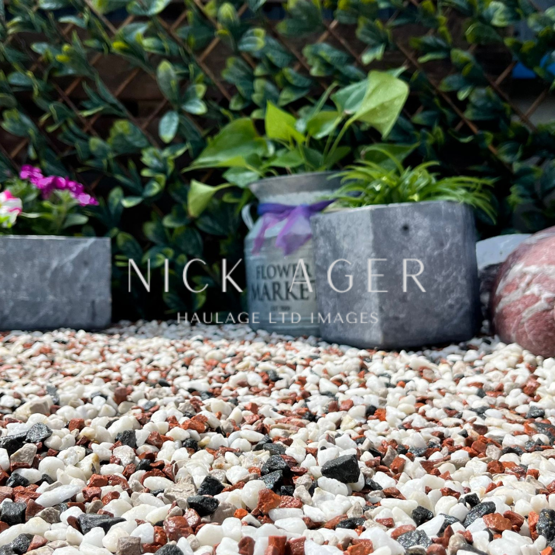 A close-up view of a garden area featuring colourful gravel, small concrete planters with flowers, and lush green foliage in the background.