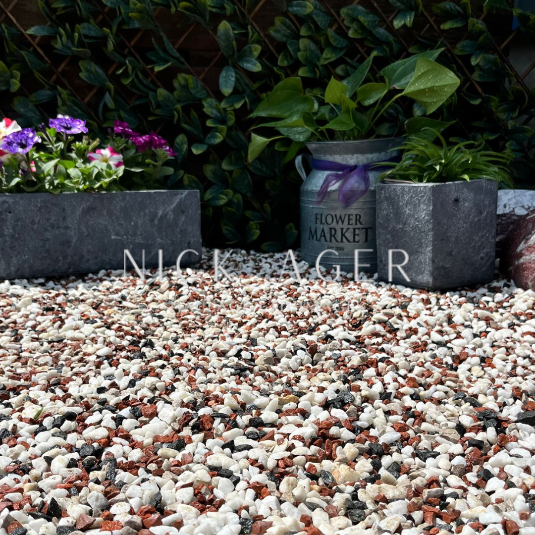 A close-up view of decorative gravel featuring a mix of white, red, and black stones, with flower pots and greenery in the background.