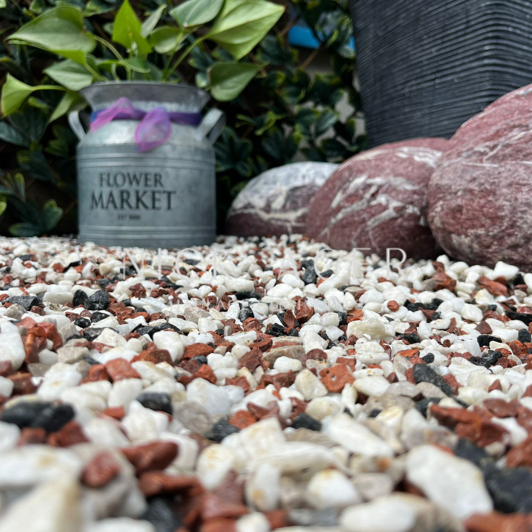 A close-up view of colourful pebbles in a garden setting, featuring a metal planter labelled 'Flower Market' and large decorative stones in the background, surrounded by green foliage.