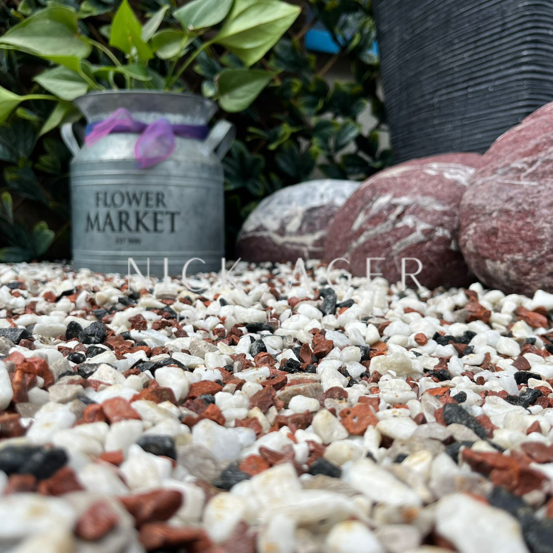 A close-up view of a garden scene with decorative gravel, a metal container labelled 'Flower Market' with a plant, and various large stones in the background.