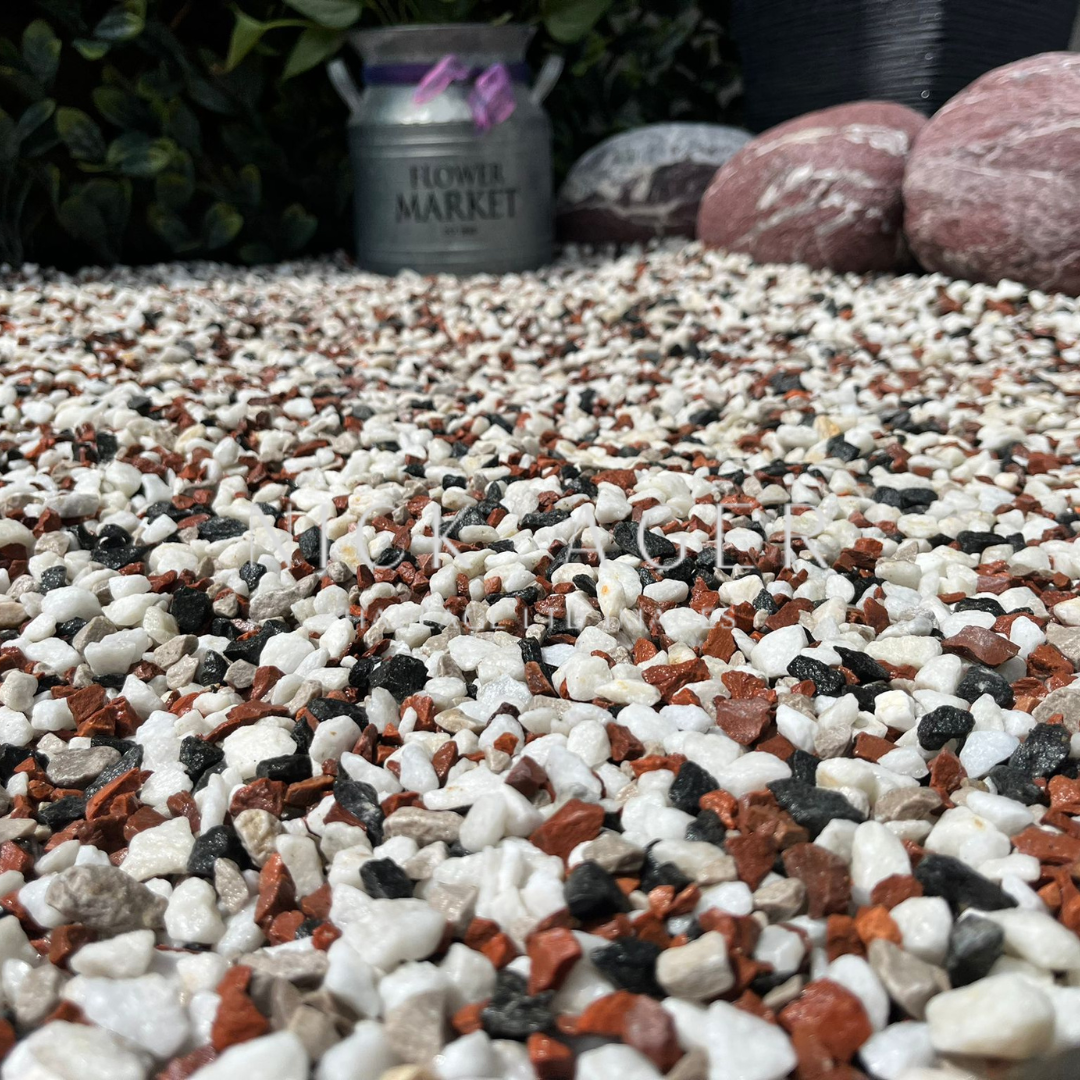 Close-up view of colourful decorative gravel composed of white, red, black, and grey stones, set against a backdrop of plants and decorative rocks.