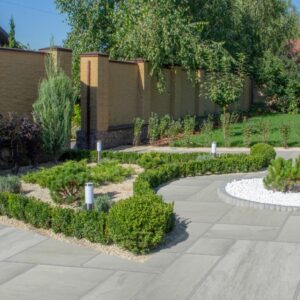 A modern garden featuring neatly trimmed hedges, decorative stones, and small trees, surrounded by a brick wall and grass lawn under clear blue skies.