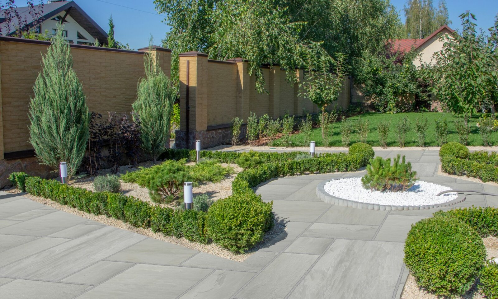 A modern garden featuring neatly trimmed hedges, decorative stones, and small trees, surrounded by a brick wall and grass lawn under clear blue skies.