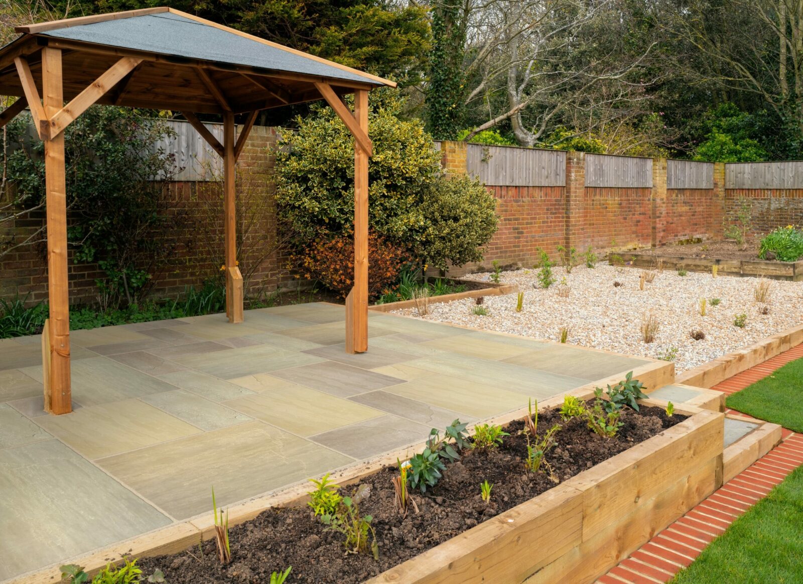 A beautifully designed garden featuring a wooden gazebo on a stone patio, surrounded by landscaped areas with plants and gravel.