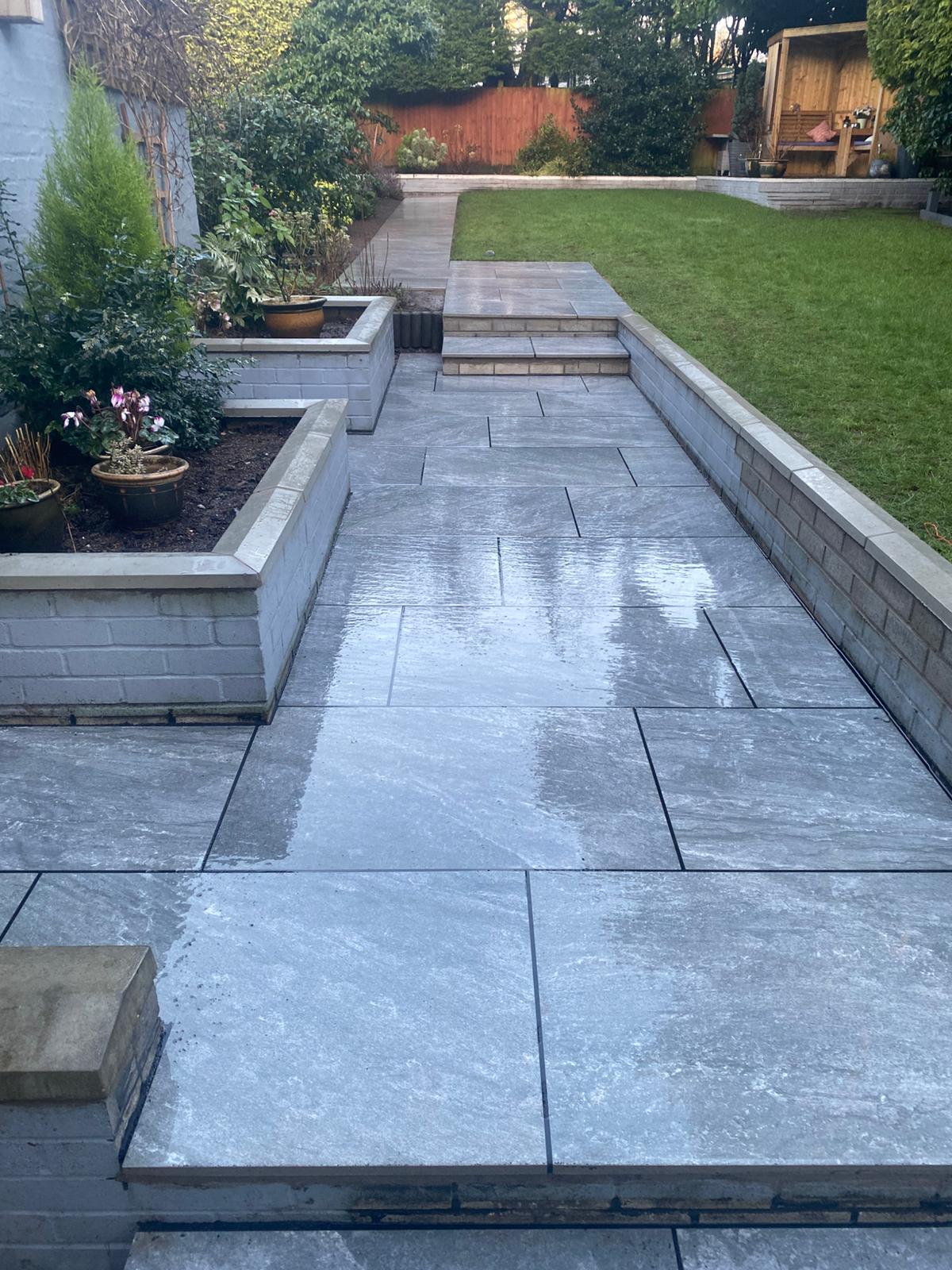 A view of a modern garden pathway made of large grey paving stones, flanked by raised planters with plants and a grassy lawn in the background.