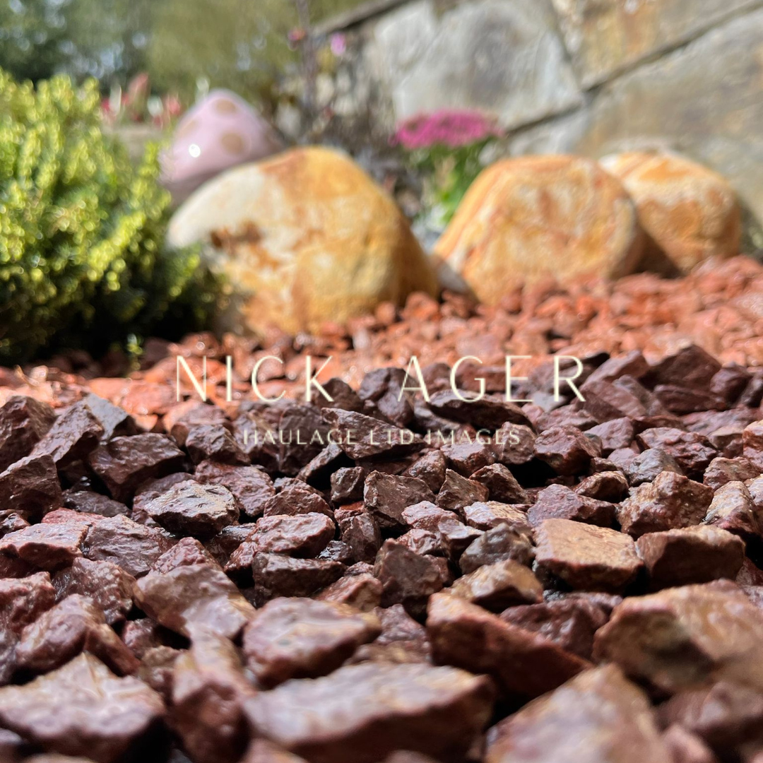 Close-up view of reddish-brown decorative gravel stones with larger stones and greenery in the background.