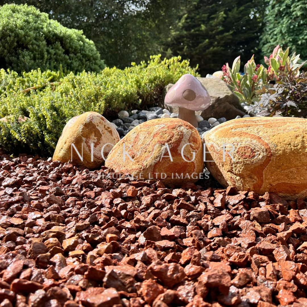 A close-up view of a garden featuring three large decorative stones on reddish gravel, surrounded by green foliage and a whimsical ceramic mushroom.