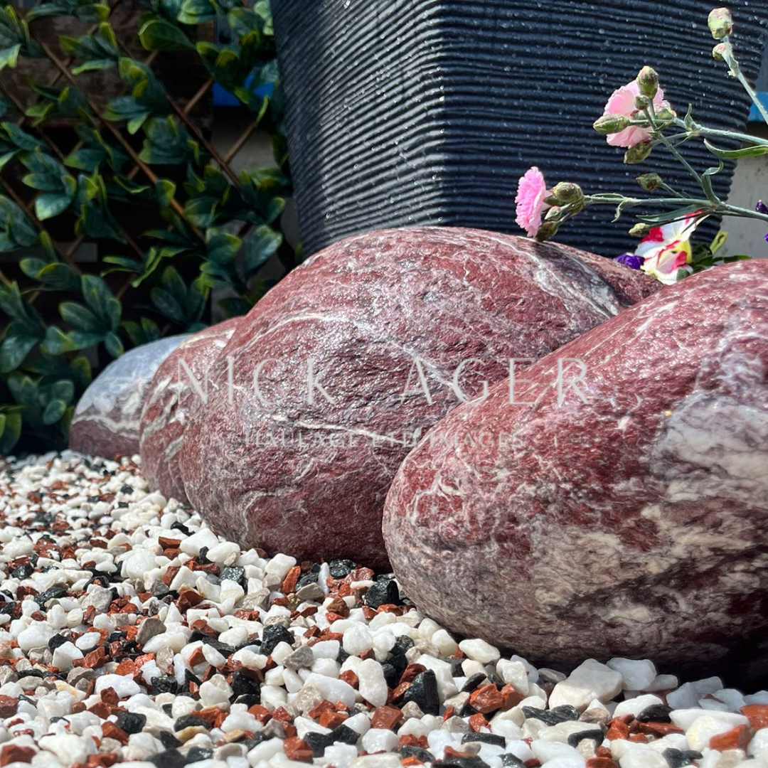 A close-up view of large decorative garden rocks sitting on a bed of multi-coloured gravel, with a black pot and flowers in the background.