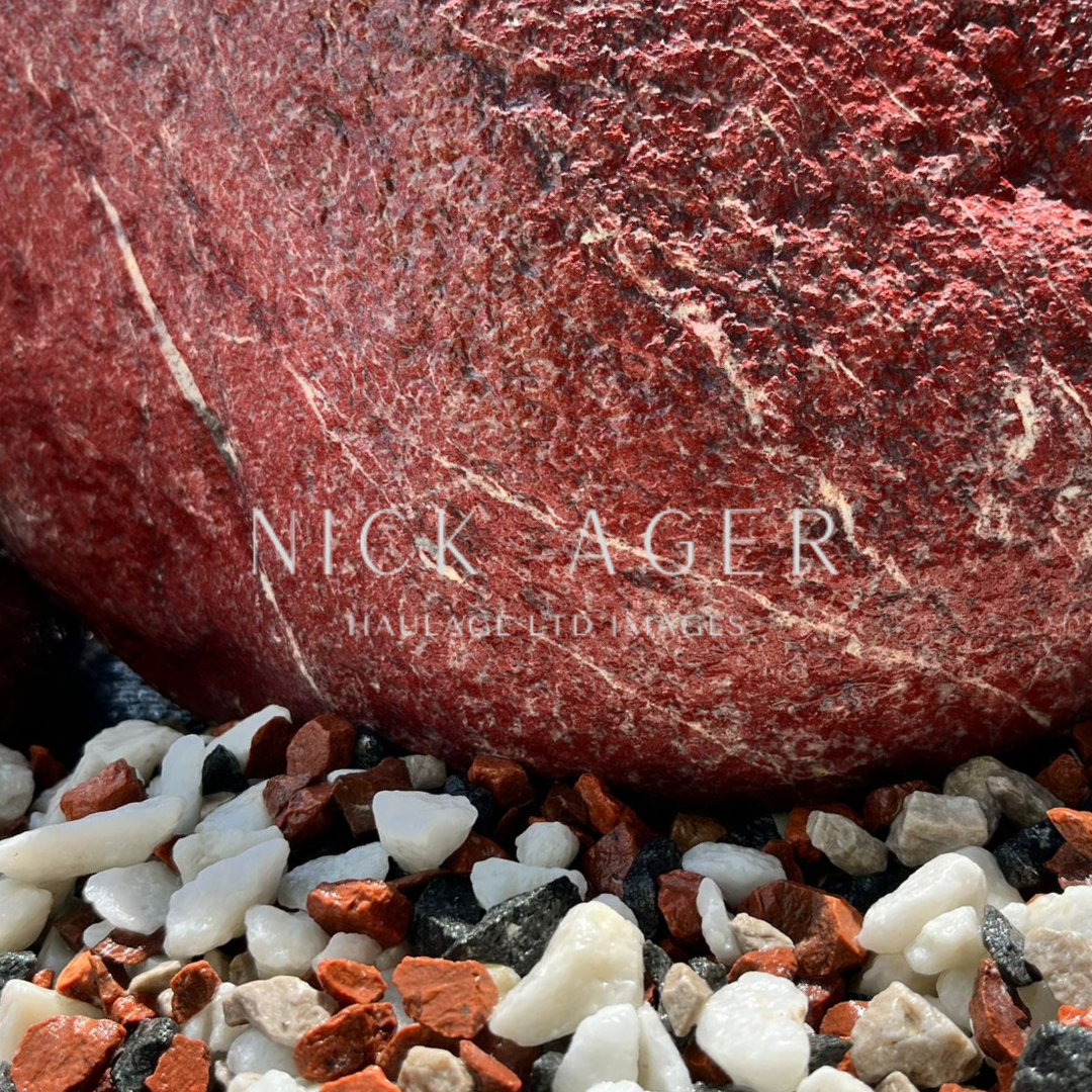 A close-up image of a large vibrant red rock with textured surface, surrounded by small white, black, and orange pebbles.