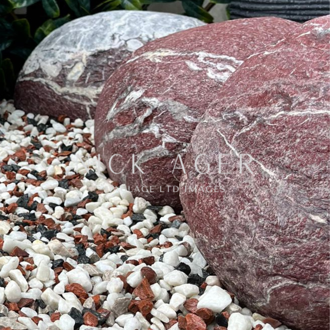 Three large decorative stones in varying shades of red and grey, surrounded by small white and red gravel, with greenery in the background.