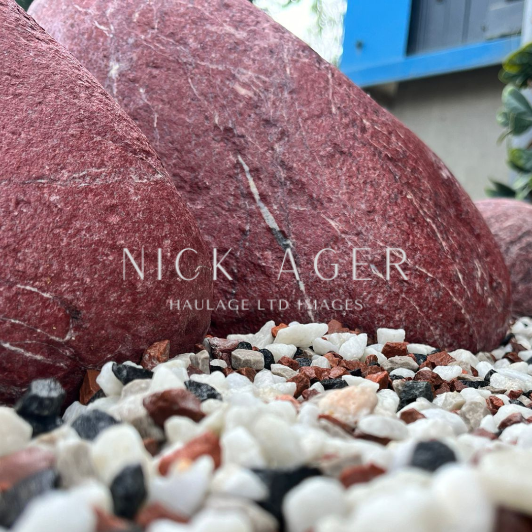 A close-up view of red decorative stones and mixed gravel including white, black, and reddish pebbles, with a blurred background.