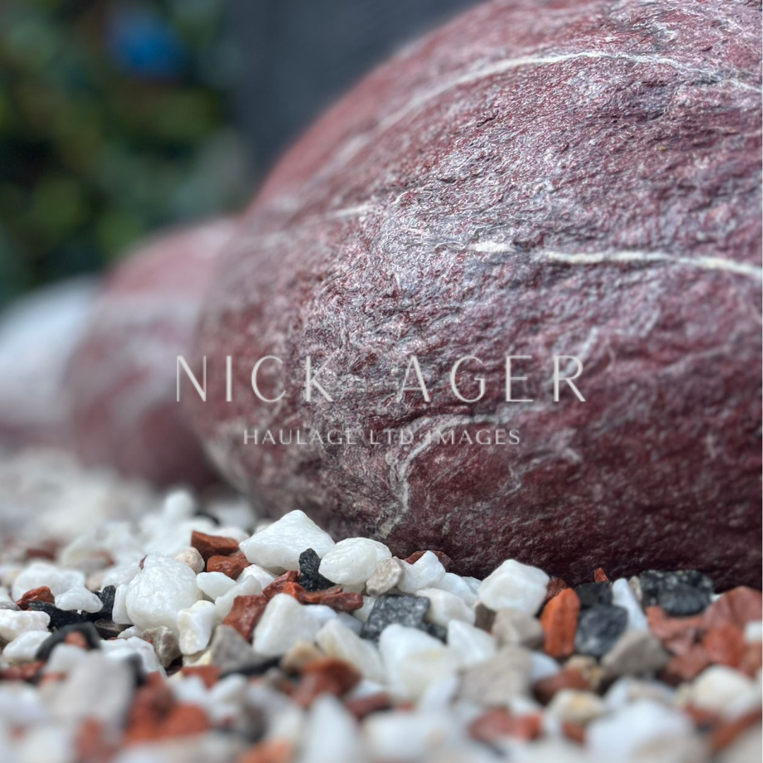 A close-up view of a large reddish stone resting on a bed of small, varying coloured pebbles, including white, black, and orange hues.