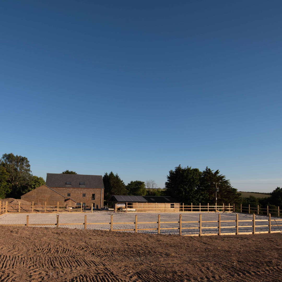 A modern equestrian facility featuring a spacious fenced riding arena, a large building with a slate roof, and lush greenery in the background under a clear blue sky.