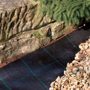 Close-up of black landscape fabric laid on soil, bordered by a stone wall and decorative gravel on one side.