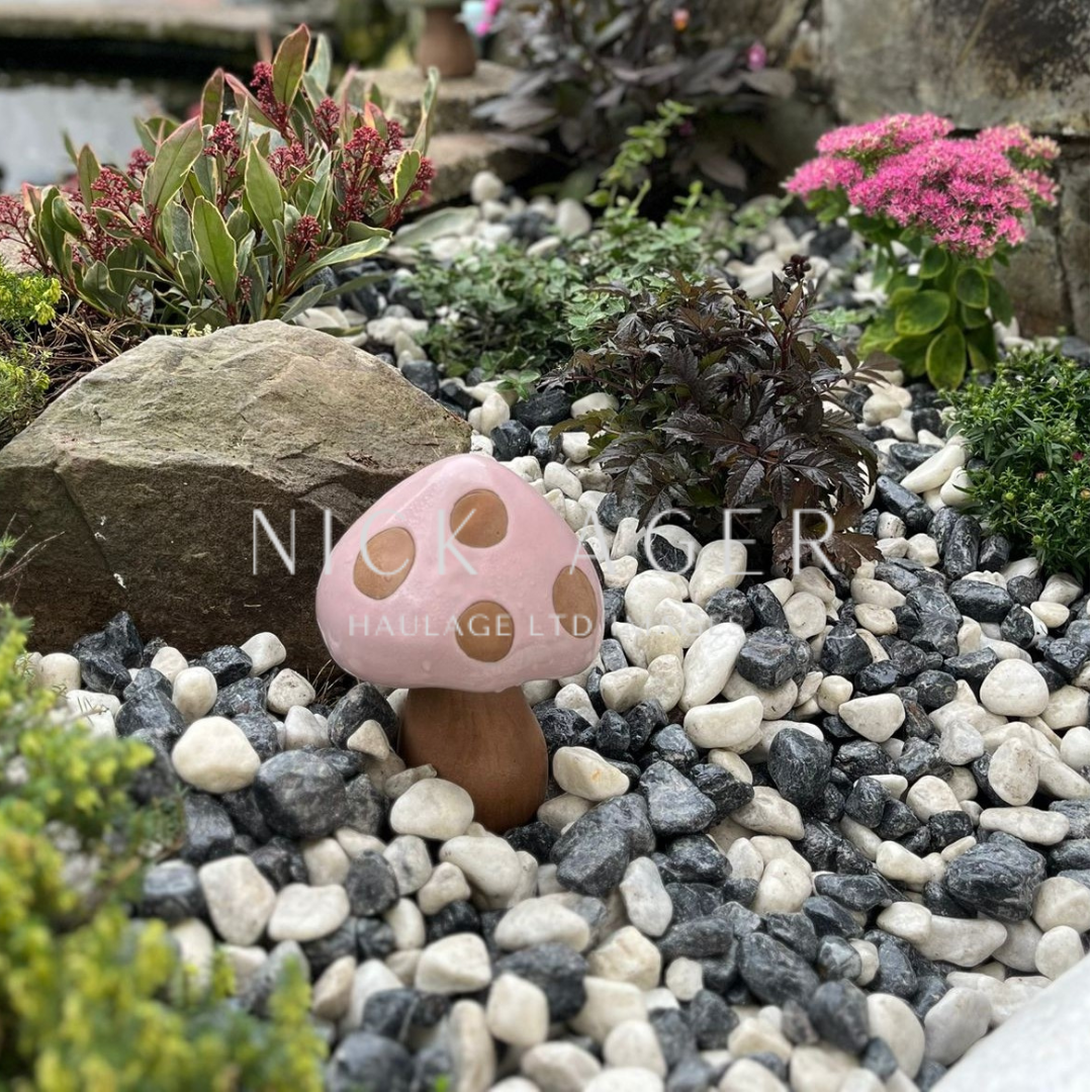 A pink decorative mushroom with brown spots stands amidst a bed of black and white pebbles in a garden setting filled with various green plants and small flowers.