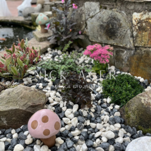 A decorative garden area featuring a pink mushroom ornament, colourful flowers, and pebbles in varying shades, surrounded by stone walls.