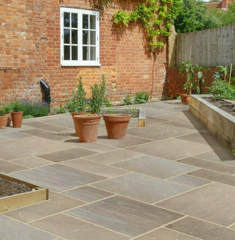 A paved garden area featuring large rectangular tiles, terracotta plant pots in the foreground, and a brick wall with a window in the background.