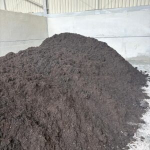 A large, dark brown pile of compost stored indoors, with metal walls and a corrugated roof in the background.
